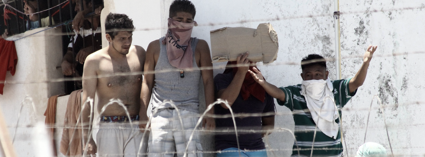 en la fotografía, personas en aparente protesta en una cárcel, con sus rostros a medio subrir aparecen detrás de un alambrado de seguridad